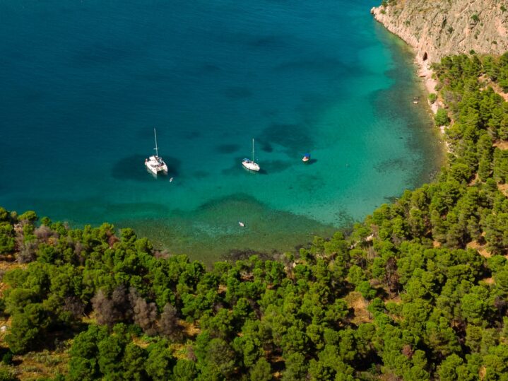 Aerial view of two sailboats anchored in clear turquoise water near a forested coastline, perfect for sailing cruises, with dense green trees covering the land and the sea gradually deepening in color.
