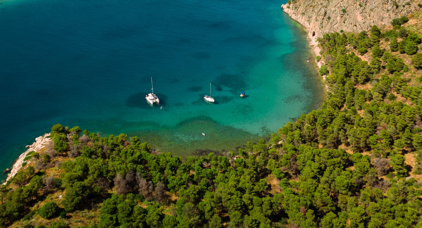 Aerial view of two sailboats anchored in clear turquoise water near a forested coastline, perfect for sailing cruises, with dense green trees covering the land and the sea gradually deepening in color.